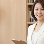 介護保険への移行（20歳前後）と入浴サービスの変化について解説する白ブラウスを着てクリップボードを持つ女性アドバイザーの横長写真