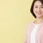 二分脊椎の入浴介助と排泄ケア（皮膚観察、導尿、褥瘡予防）を優しく解説するピンクのカーディガンを着た女性アドバイザーの横長写真